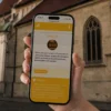 Hand holding a smartphone showing a treasure-hunt app screen titled 'Stopa 13' against a historic stone church backdrop
